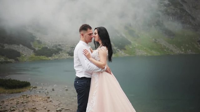 Portrait lovely wedding couple stands near lake in mountains, hugging kissing on a cloudy day groom happy beautiful dress celebration party face bridge summer young nature outdoor