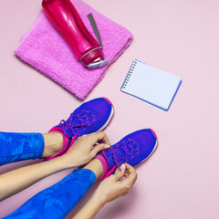 Young woman in sporting blue leggings laces sneakers, preparing for training. Accessories for sports, bottle of water towel notebook on pastel pink background flat lay top view. Fitness concept