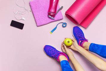 Young woman in blue sneakers holds apple in his hands, preparing for training. Bottle of water, yoga mat, phone, headphones on pastel pink background flat lay top view. Sports shoes, Fitness concept