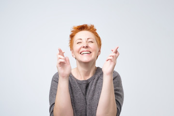 Happy matureredhead female eager to win competition, keeps fingers crossed as waits for results, smiles broadly, shows white perfect teeth, poses in studio.