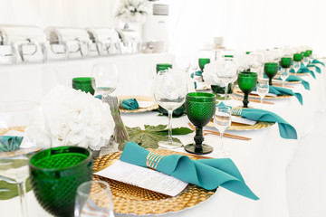 Interior of tent for wedding diner, ready for guests. Served round banquet table outdoor in marquee decorated hydrangea flowers, Golden dishes and green napkins. Catering concept