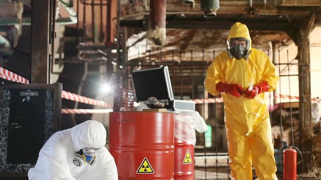 Two scientists wearing white and yellow hazmat suits working at old nuclear power station, using special devices. Radiation. Indoors. Dangerous work.