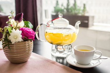 Glass teapot with hot pineapple tea with pineapple slices in a cafe. A cup of fragrant yellow tea, a glass teapot and beautiful pink flowers in a vase.