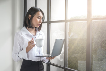 Young Asian business woman using her tablet PC while standing relaxed near window at her office