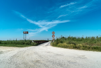 Schotterweg über eine alte eiserne Brücke in die weite Ebene mit Wegweiser nach Valli di...