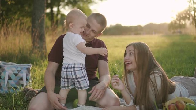 Young man, woman and first child have good weekend in nature park, American dad and mom are playing with cute baby, having fun with smiles, sitting on green grass on sunny day. Concept: family