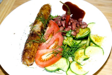 On a white plate with a serving option lies a delicious meal. Spices are sprinkled with fresh cucumbers, red beans, tomatoes, dill and fried in a skillet, a natural sausage, which is decorated with ch