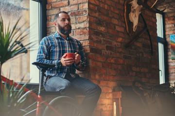 Handsome bearded hipster male in a blue fleece shirt and jeans holds a cup of morning coffee while sitting on a window sill at a studio with a loft interior.