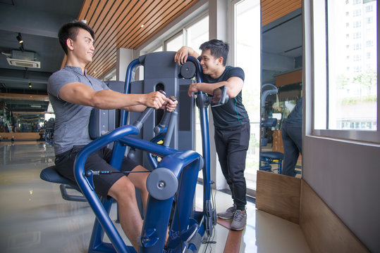 Asian Man Training On Exercise Machine And Talking With Friend. Smiling Young Guys In Gym. Bodybuilding Concept.