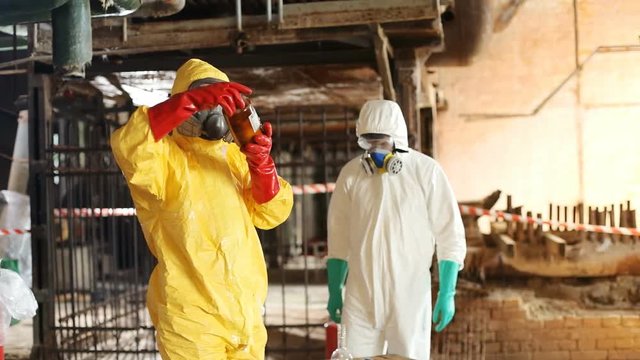 Scientists working at old factory after accident with dangerous gas. Guy in yellow hazmat suit considering carefully jar with strange liquid.