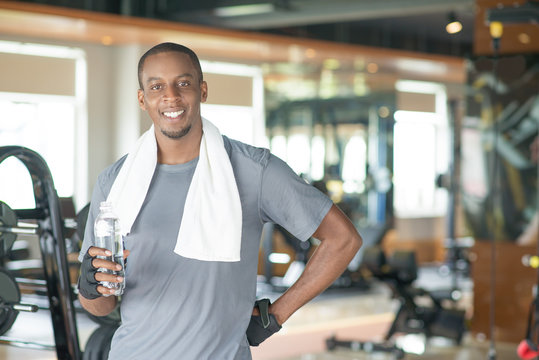 Happy Black Man Holding Water Bottle, Standing In Gym And Looking At Camera. Young Guy Wearing Towel Around Neck And Having Rest. Workout And Break Concept. Front View.