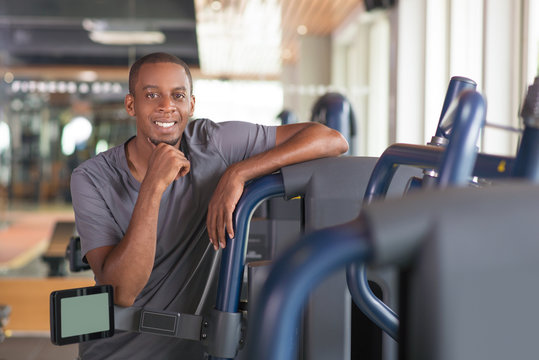 Relaxed Black Man Leaning On Gym Equipment And Looking At Camera. Smiling Young Guy Wearing T-shirt, Standing And Having Rest. Workout And Break Concept. Front View.