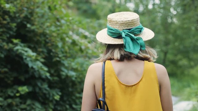 Young woman wearing romantic hat walking through green forest. Women’s back. Feminine image. Summertime. Outdoors. Warm weather.