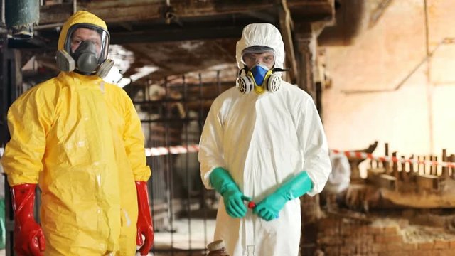 Two smart Caucasian scientists wearing hazmat outfits working at abandoned factory. Guys looking attentively at camera. Indoors. Invisible danger.