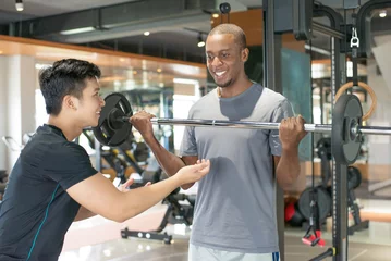 Schilderijen op glas Smiling black man lifting barbell with personal trainer. Young guy training with gym equipment in background. Bodybuilding concept. © Mangostar