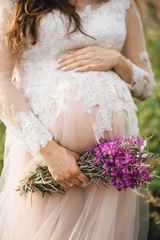 Close-up of unrecognizable pregnant woman with hands over tummy in a white lace negligee and flowers