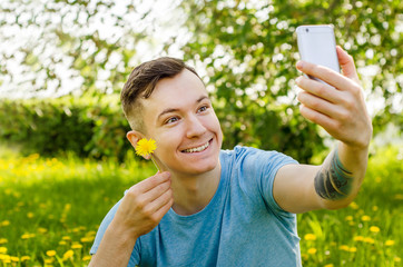 Young smiling guy sits on a green grass with dandelions and does selfie on a smartphone and holds flower of dandelion.