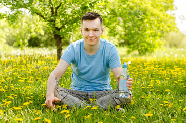 Naklejka na ściany i meble Young guy dressed in a blue t-shirt holds bottle of water, sits on a green grass and yellow dandelion background.
