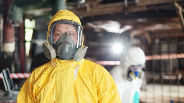 Young Caucasian man wearing new yellow hazmat outfit looking at camera in background working colleague. Close-up. Dangerous work.