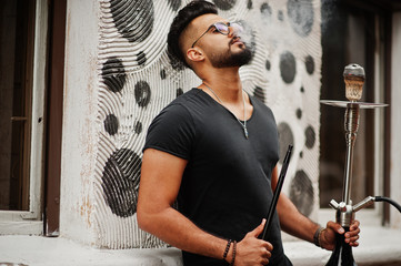 Stylish beard arabian man in glasses and black t-shirt smoking hookah outdoor. Arab model having rest.