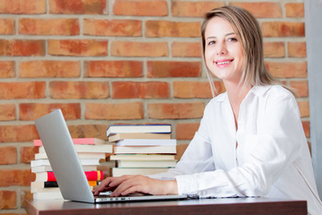 Young businesswoman sitting on a table with laptop computer and books. Brick wall on background