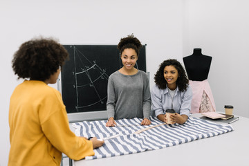 Creative team of smiling curly African American women planning new startup project in workplace. Brainstorming. University students learning fashion design at atelier. Young hipsters working together.
