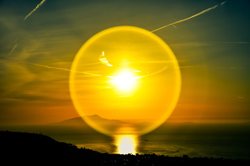 Efecto circular de luz del sol sobre el lente durante un atardecer en las costas de Sorrento al sur de Italia. Cielo de tonos naranjas y azules y monte Vesubio de fondo. 