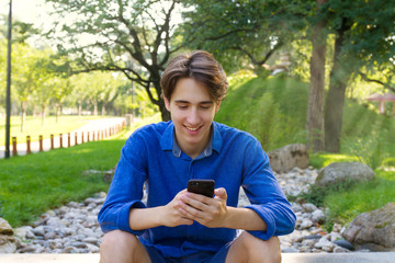 Outside portrait of young man in park. Smiling handsome teenager typing or reading at phone