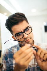 Handsome young man choosing eyeglasses frame in optical store.
