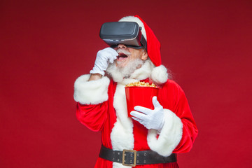 Christmas. Santa Claus in black virtual reality glasses holding a red bucket of popcorn. Watching a movie in virtual reality. Isolated on red background.