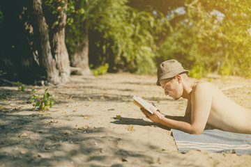 young handsome attractive man in panama lying on sandy heaven beach and reading a book on a summer day