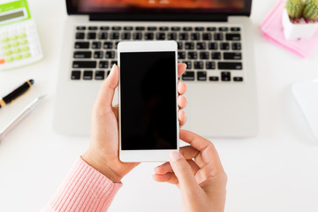 Woman hand holding white mobile phone on a table with a laptop in office.