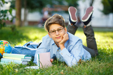 cute, young boy in round glasses and blue shirt reads book lying on the grass in the park. Education, back to school concept