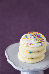 Three frosted sugar cookies, white frosting, colored sprinkles stacked on a white cake plate on a purple background
