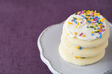 Three frosted sugar cookies, white frosting, colored sprinkles stacked on a white cake plate on a purple background
