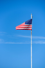 Fototapeta premium American flag on top white flagpole, flying in the breeze, blue sky background, wispy white cloud, stars and stripes, red, white, and blue 