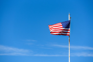 American flag on top white flagpole, flying in the breeze, blue sky background, wispy white cloud, stars and stripes, red, white, and blue
