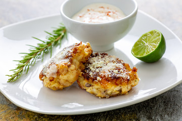 Two shrimp cakes on a white plate with a lemon garlic dipping sauces lemon wedge and a stem or rosemary on a wooden table.