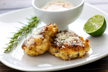 Two shrimp cakes on a white plate with a lemon garlic dipping sauces lemon wedge and a stem or rosemary on a wooden table.