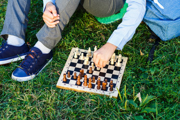 cute boy in riound glasses and blue shirt sits on the grass in the park and plays chess at wooden...
