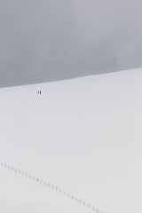 A very minimalistic view of two distant people over a mountain covered by snow, near a fence