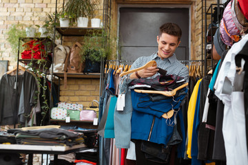 Cheer clothes. Cheerful emotional handsome man smiling and feeling satisfied with his weekend shopping while holding great amount of clothes on hangers