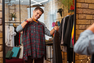 Perfect shirt. Cheerful emotional young man feeling satisfied with new beautiful clothes while visiting a modern clothes shop and looking at his reflection in the mirror