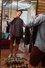 Smiling buyer. Cheerful emotional young man smiling and trying on fashionable shirts while standing in front of the mirror
