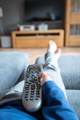 Eine kindliche Hand hält eine moderne Fernbedienung und zeigt damit auf den Fernseher. Die Füße liegen auf der Couch