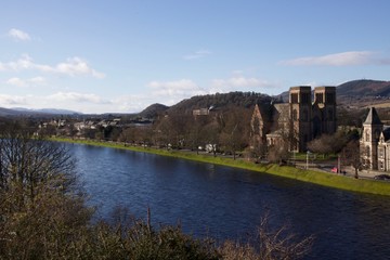 Fototapeta na wymiar A cathedral in Inverness runs along the Scottish city river on a sunny day with blue skies and green trees in the foreground