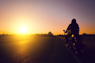 Fototapeta na wymiar Silhouette of biker woman with his motorbike(motorcycle ) on street,he shoulder backpack, enjoying freedom and active lifestyle, having fun on a bikers tour sunset background.