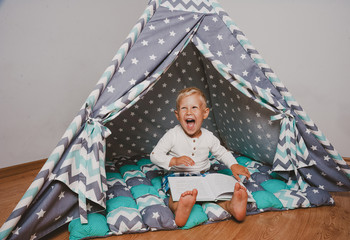 Happy boy smiling and holding book sitting in wigwam tent in the room © Vulp