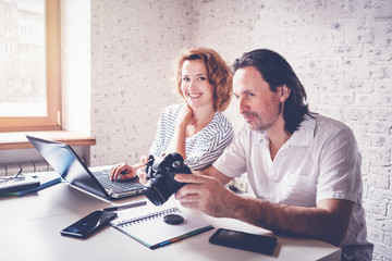 A middle-aged man and a young woman are sitting at a table with a laptop, camera and diary. Training and master classes in photography and processing, education concept, creative professions