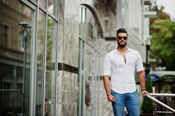 Stylish tall arabian man model in white shirt, jeans and sunglasses posed at street of city. Beard attractive arab guy.